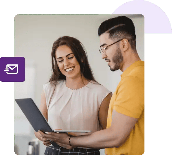 A man holds his laptop and shows his screen to a woman next to him that’s smiling.
